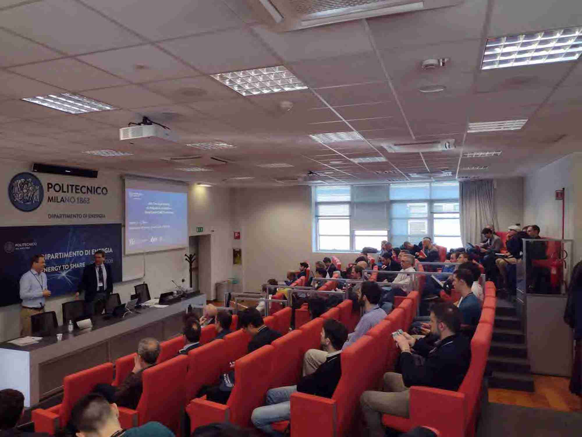 Foto di un aula conferenze con sedie rosse sulla destra. Sulla sinistra persone che parlano