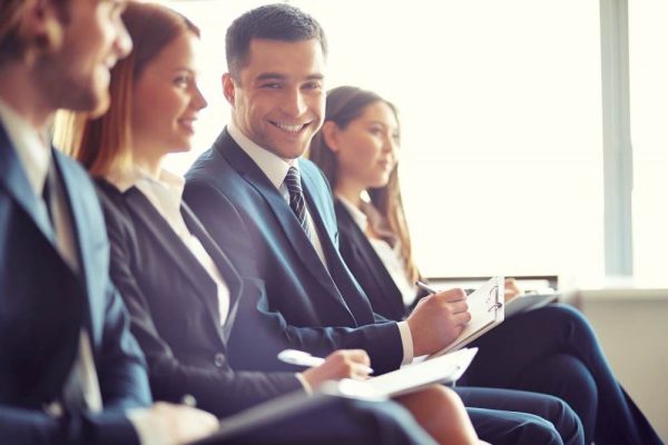 Uomo in abito d'affari sorride a favore di telecamera in mezzo a colleghi che prendono appunti
